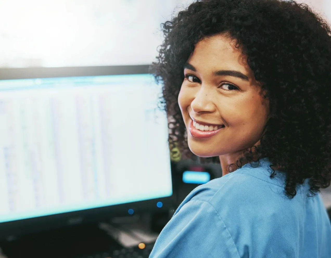 Smiling woman in blue scrubs working at a computer, representing Medical Office Administration Certificate Program skills.