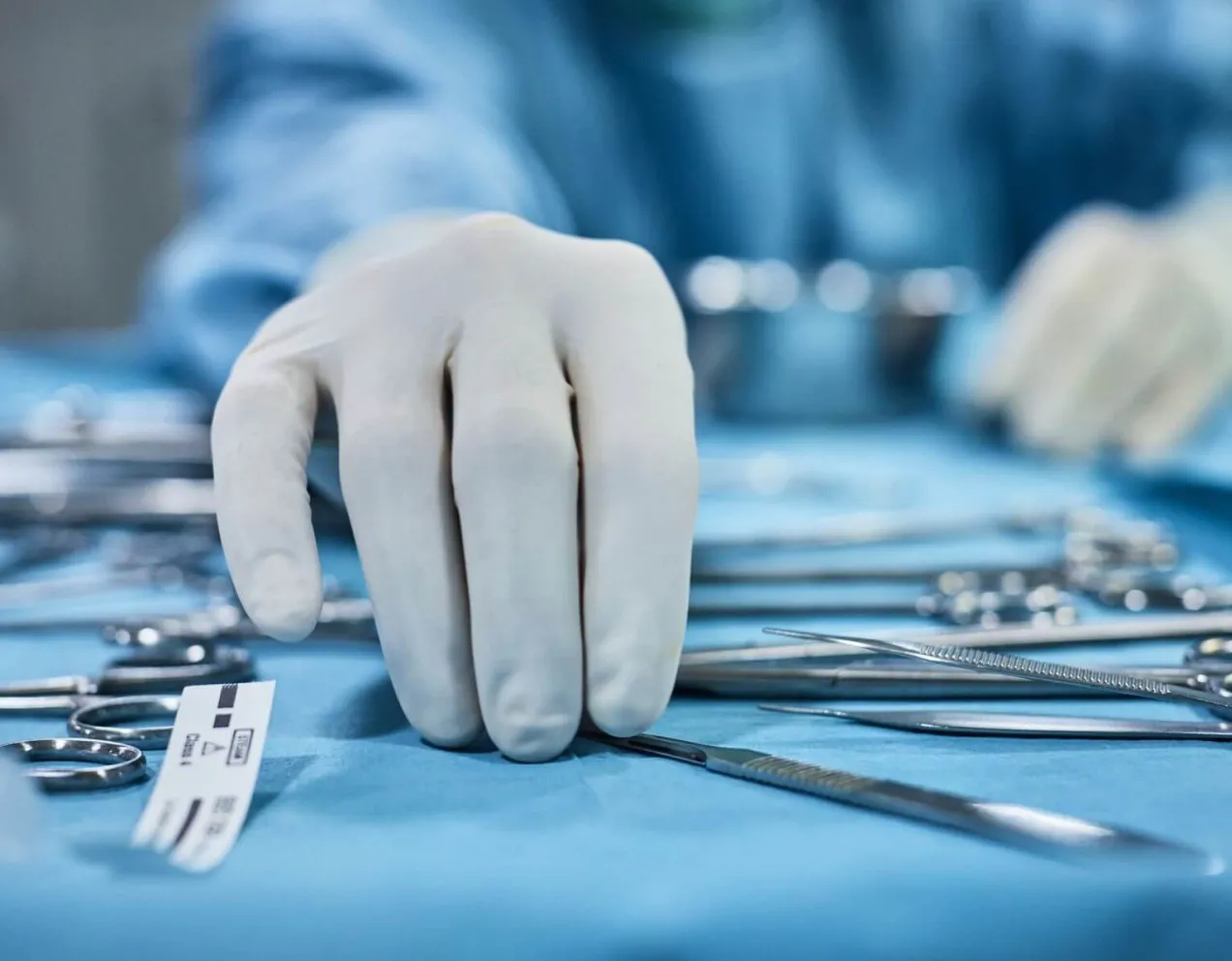 A close-up view of a gloved hand, representing a surgical technologist reaching for a scalpel among a spread of various sterile surgical instruments on a blue instrument tray.