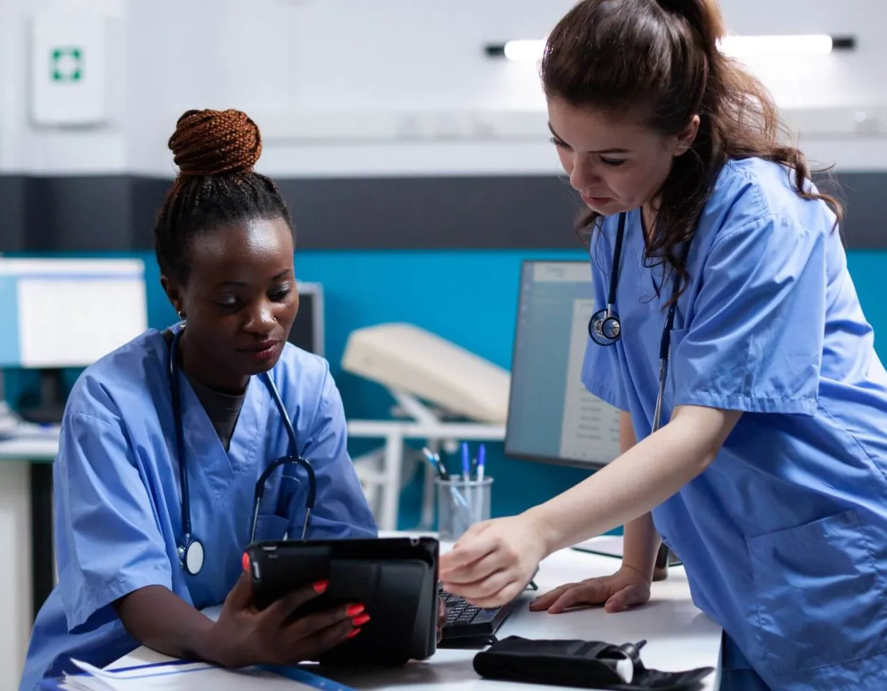 A medical assistant and a nurse in blue scrubs collaborate in a modern clinic, reviewing patient data on a digital tablet to illustrate the differences between their clinical and administrative roles.