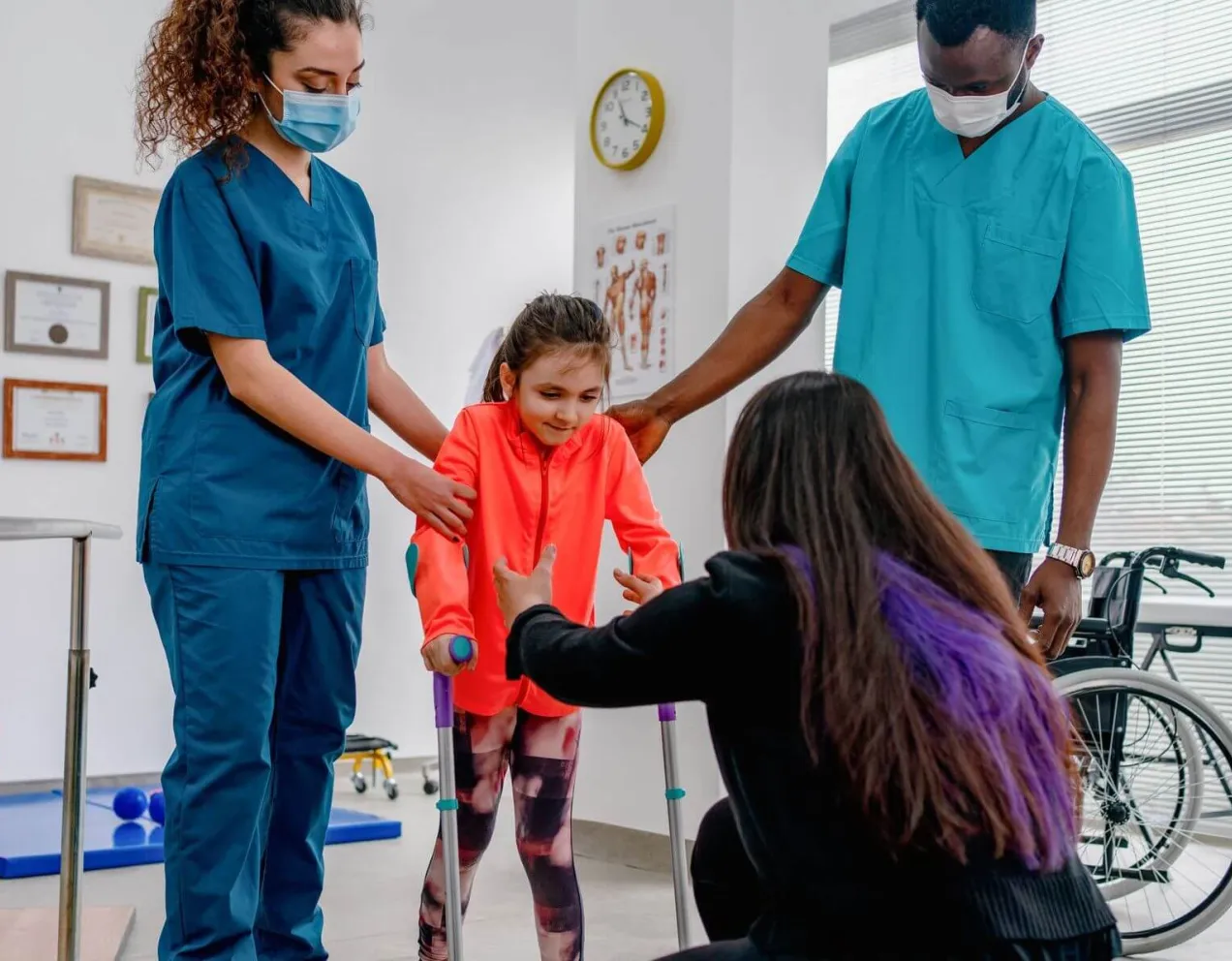 A pediatric physical therapist assistant supports a young girl during a therapeutic exercise session in a brightly colored clinic.