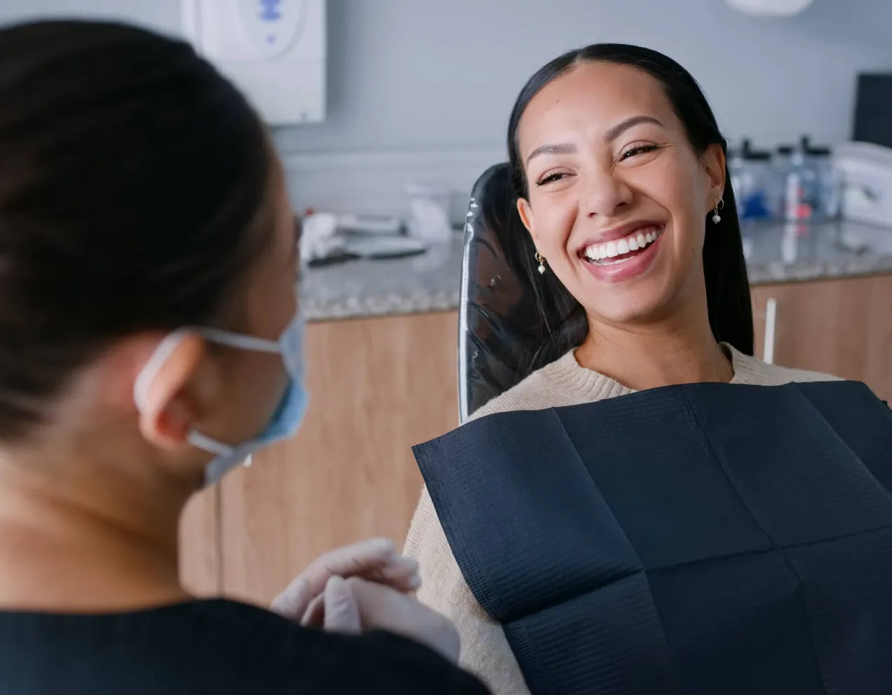A smiling dental hygienist wearing a face mask and gloves demonstrates excellent communication skills while speaking with a happy patient in a dental chair.