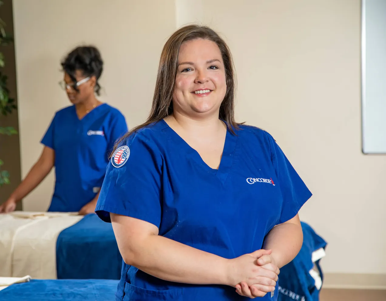 A smiling Concorde massage therapy student wearing blue scrubs with a military-themed patch stands in a healthcare training lab, illustrating how to use military tuition assistance for career training.