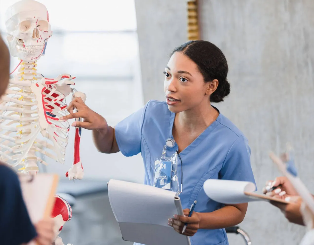 A female nurse educator uses a human skeleton model to teach clinical anatomy to a group of nursing students.