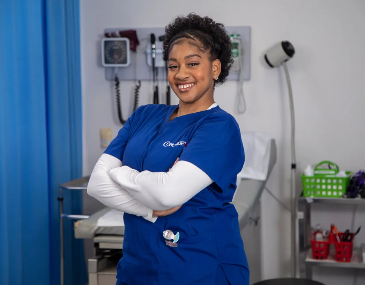 A female medical assistant wearing blue scrubs stands with arms crossed and smiling in a hospital setting