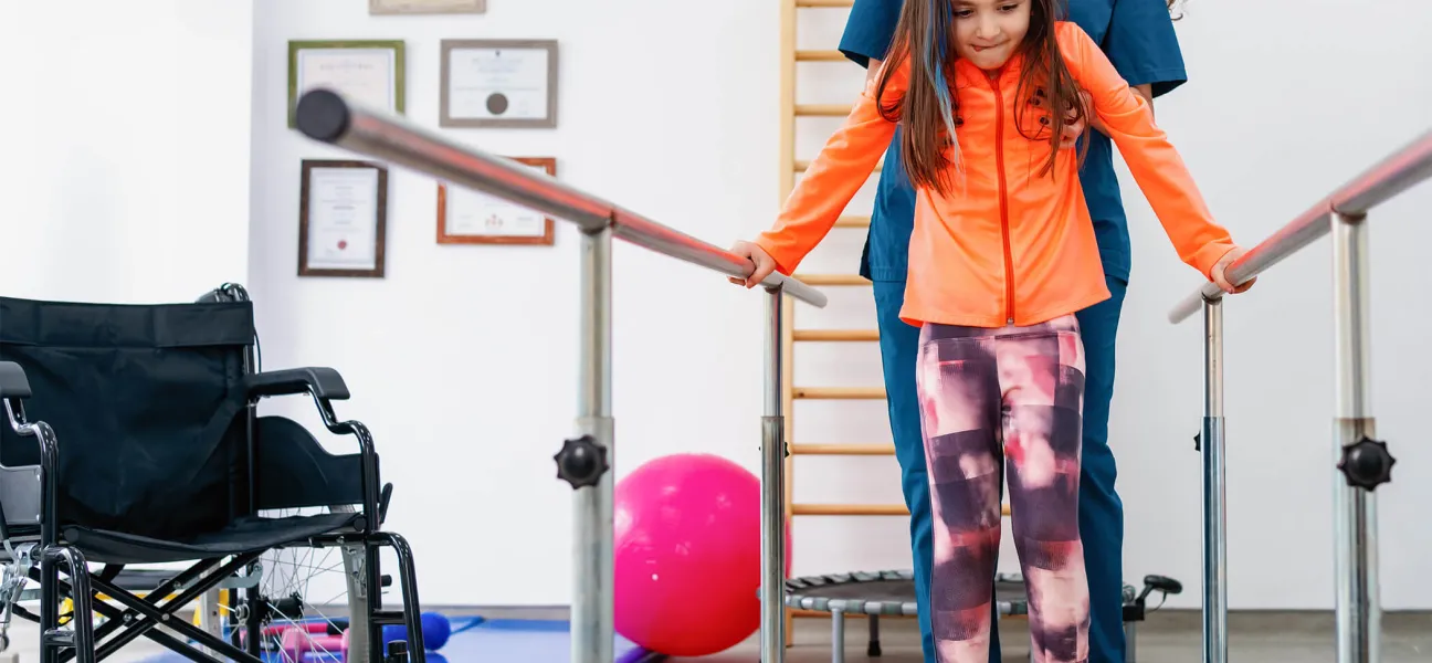 Occupational therapy assistant helps young girl improve mobility using parallel bars during rehabilitation.
