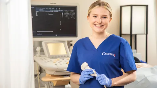 Smiling sonography student in scrubs holding ultrasound probe in clinical training setting.