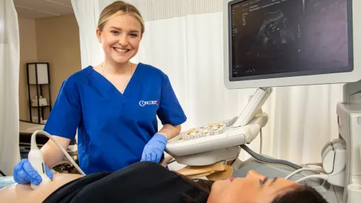 Sonography student performing ultrasound exam on patient in lab with imaging screen visible.