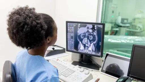 Radiologic technologist analyzing abdominal CT scan on monitor in a medical imaging control room.
