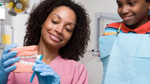Dental assistant showing a child how to brush teeth using a model, demonstrating oral hygiene education in a dental care setting.