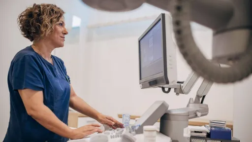 A female sonographer in blue scrubs is professionally operating an ultrasound machine, reviewing the patient data on the connected medical monitor.