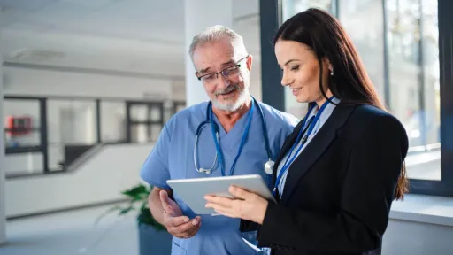 A healthcare administrator in a business suit and a senior doctor in scrubs review data on a tablet, illustrating a typical administrative discussion in a modern medical setting.
