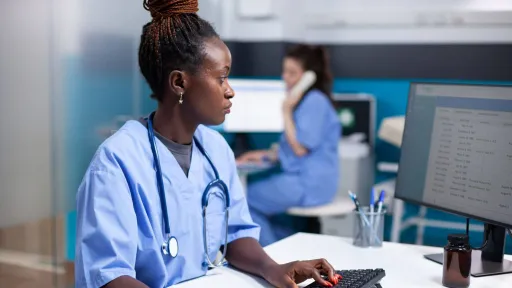A medical assistant wearing scrubs and a stethoscope sits at a desk, focused on entering patient data into an Electronic Health Record (EHR) system on a computer.