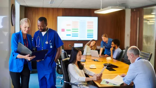 Diverse group of healthcare professionals and medical administrative staff in a meeting room, reviewing documents and screen data.