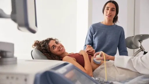 A smiling pregnant woman lies on an examination table getting an ultrasound, while her adult daughter stands by and holds her hand in a warm, supportive medical moment.