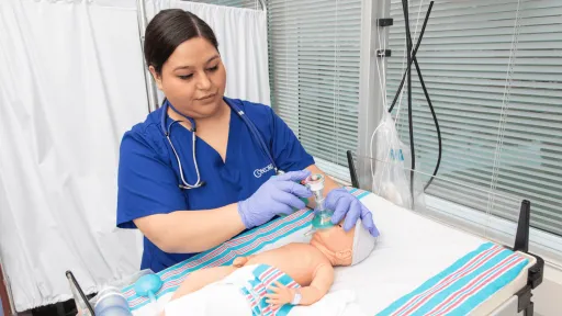 A female respiratory therapist with dark hair wearing blue scrubs assisting an infant patient with breathing in the hospital