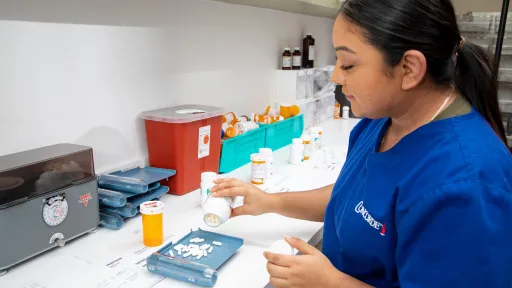 A female pharmacy technician in a blue uniform is accurately counting prescription pills using a counting tray on a clean white workbench.