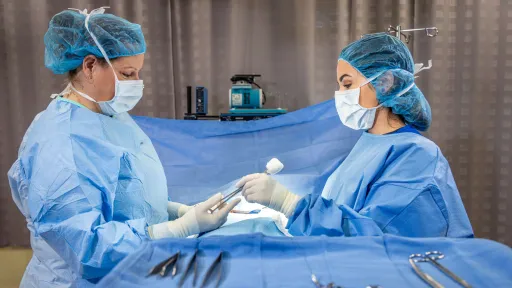 A female surgical technologist in face mask and surgical cap and gown passing surgical instrument to female surgeon in operating room