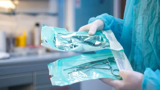 A sterile processing technician wearing a blue surgical gown and white gloves holds up two heat-sealed pouches containing sterilized surgical instruments.