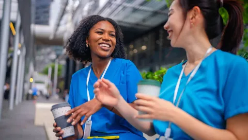 Two diverse female nurses in blue scrubs laugh and enjoy a coffee break together outside, highlighting the social and supportive environment of the nursing profession.