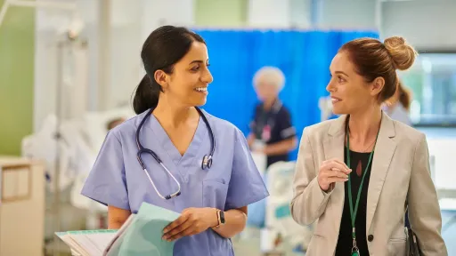 A smiling healthcare administrator in a grey blazer walks through a hospital ward while discussing patient care with a nurse in blue scrubs.
