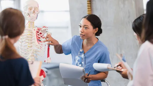 A nurse educator points to the rib cage of a human skeleton model while teaching a group of nursing students in a clinical classroom setting.