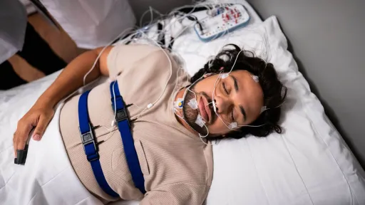A patient lies in a bed with electrodes attached to his head and face while wearing chest belts for a polysomnography test.