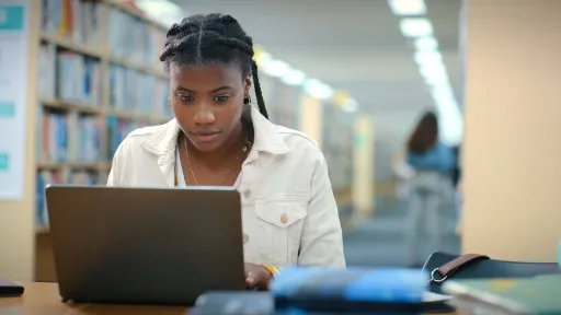 A focused nursing student working on her laptop in a quiet library surrounded by bookshelves while preparing for the NCLEX.