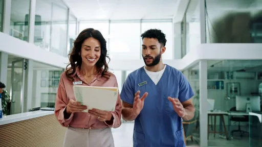 A female nurse leader in professional attire discusses patient documentation with a male colleague in a modern healthcare facility.