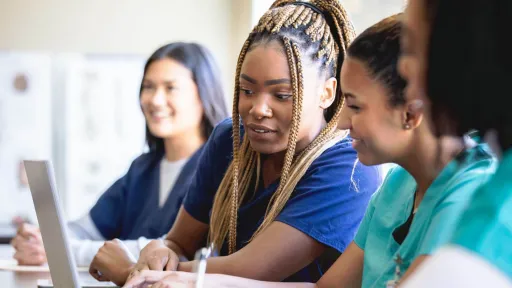 A diverse group of healthcare students in scrubs look at a laptop screen together in a classroom during their transition training to become registered nurses.