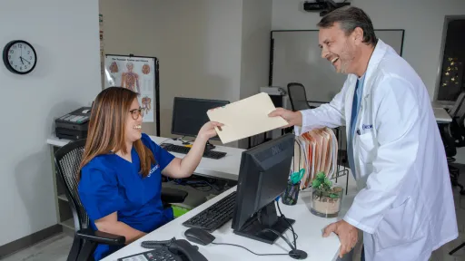 Female medical office assistant in blue scrubs sitting at front desk in medical office hands a patient file to a male doctor wearing white coat