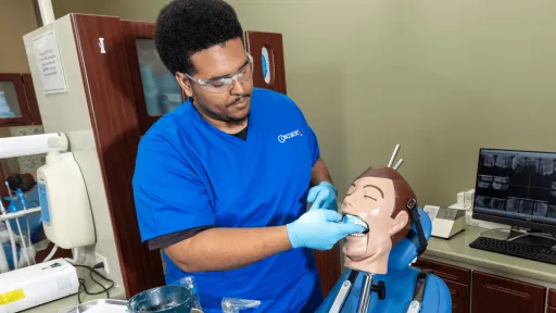 Male dental assistant with dark hair and blue scrubs works on dummy in lab setting to create dental impressions for dental work