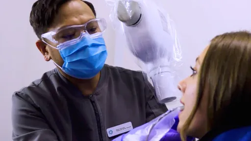 A male dental assistant in grey scrubs works with patient in dentist's chair