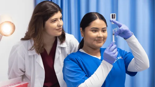 A female medical assistant wearing blue scrubs fills a syringe with medicine while a female physician supervises