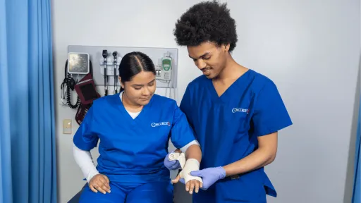A male medical assistant wearing blue scrubs wraps gauze bandage around a female medical assistant's wrist