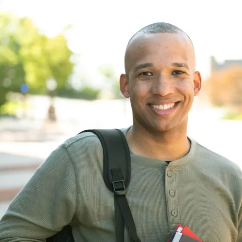 A smiling male military veteran student stands on a sunny college campus wearing a backpack and holding notebooks.