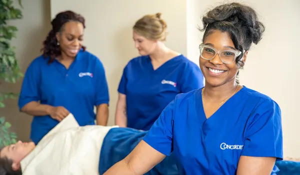 A massage therapist leans on her table and smiles at the camera.