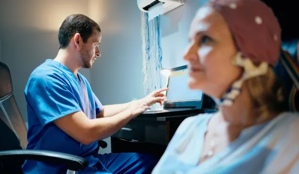 EEG technician monitoring brain wave activity on screen while patient undergoes neurological testing in clinical setting
