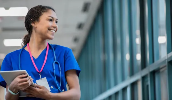 Smiling travel nurse in scrubs holding a tablet in a hospital hallway, representing confidence and readiness for a traveling nurse career