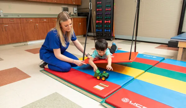 Occupational therapy assistant guiding a child through sensory motor play using colorful mats and a toy tractor during a pediatric therapy session.