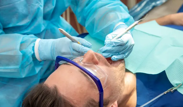 A close-up view of a dental hygienist wearing a blue gown and gloves using dental tools to work on a male patient who is wearing protective eyewear.