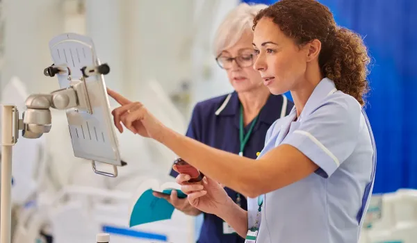 Two female nurses in professional uniforms review medical data on a wall-mounted digital terminal while one holds a medication bottle and a reference book.