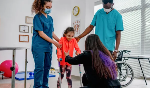 A pediatric physical therapist assistant supports a young girl during a therapeutic exercise session in a brightly colored clinic.