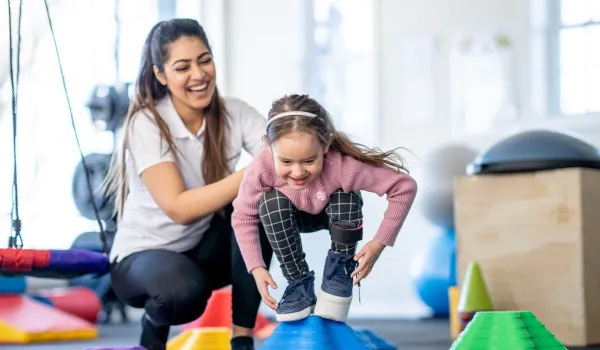 A young child balances on a large blue therapy ball while being guided through stability exercises by a pediatric physical therapist assistant.