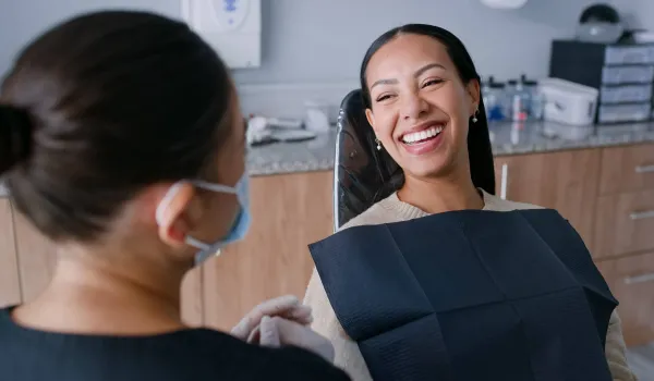 A smiling dental hygienist wearing a face mask and gloves demonstrates excellent communication skills while speaking with a happy patient in a dental chair.
