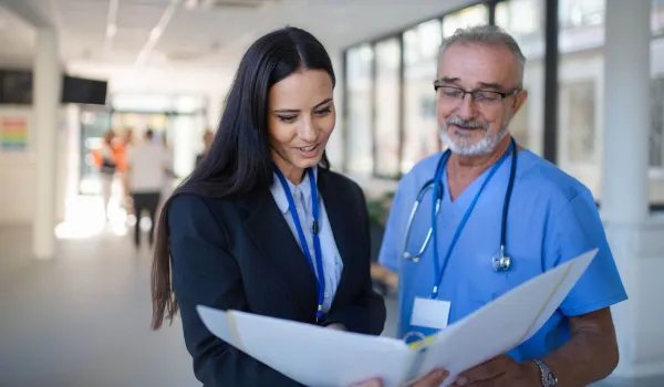 A female healthcare administrator in a black suit reviews medical documents in a binder with a senior male doctor in blue scrubs.