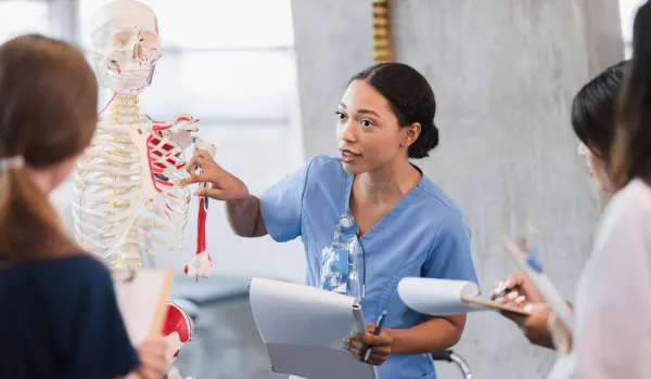 A female nurse educator uses a human skeleton model to teach clinical anatomy to a group of nursing students.