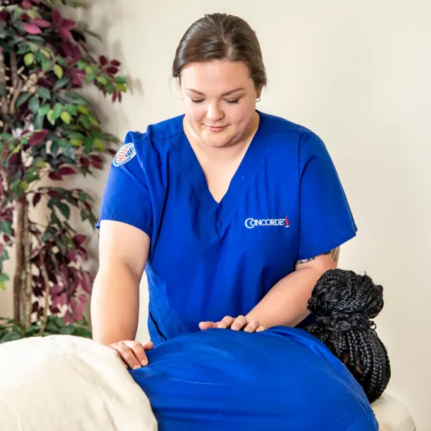 A professional massage therapist wearing blue scrubs with a Concorde logo is performing a leg massage on a client in a bright treatment room.