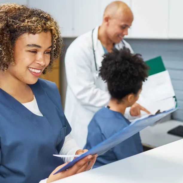 A smiling medical assistant in blue scrubs stands at a reception counter reviewing a patient chart while a doctor and another medical assistant discuss data on a computer screen in the background.