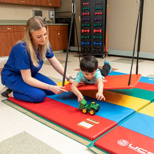 Occupational therapy assistant guiding a child through sensory motor play using colorful mats and a toy tractor during a pediatric therapy session.
