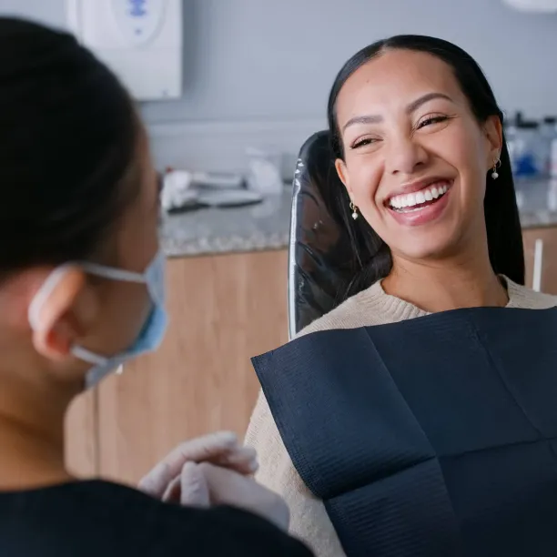 A smiling dental hygienist wearing a face mask and gloves demonstrates excellent communication skills while speaking with a happy patient in a dental chair.